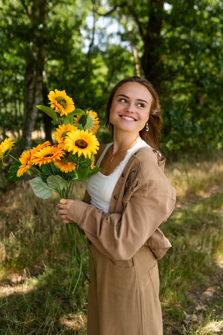 Vrouw in een beige jas en witte top die een bos zonnebloemen vasthoudt in een zonnige, groene omgeving.