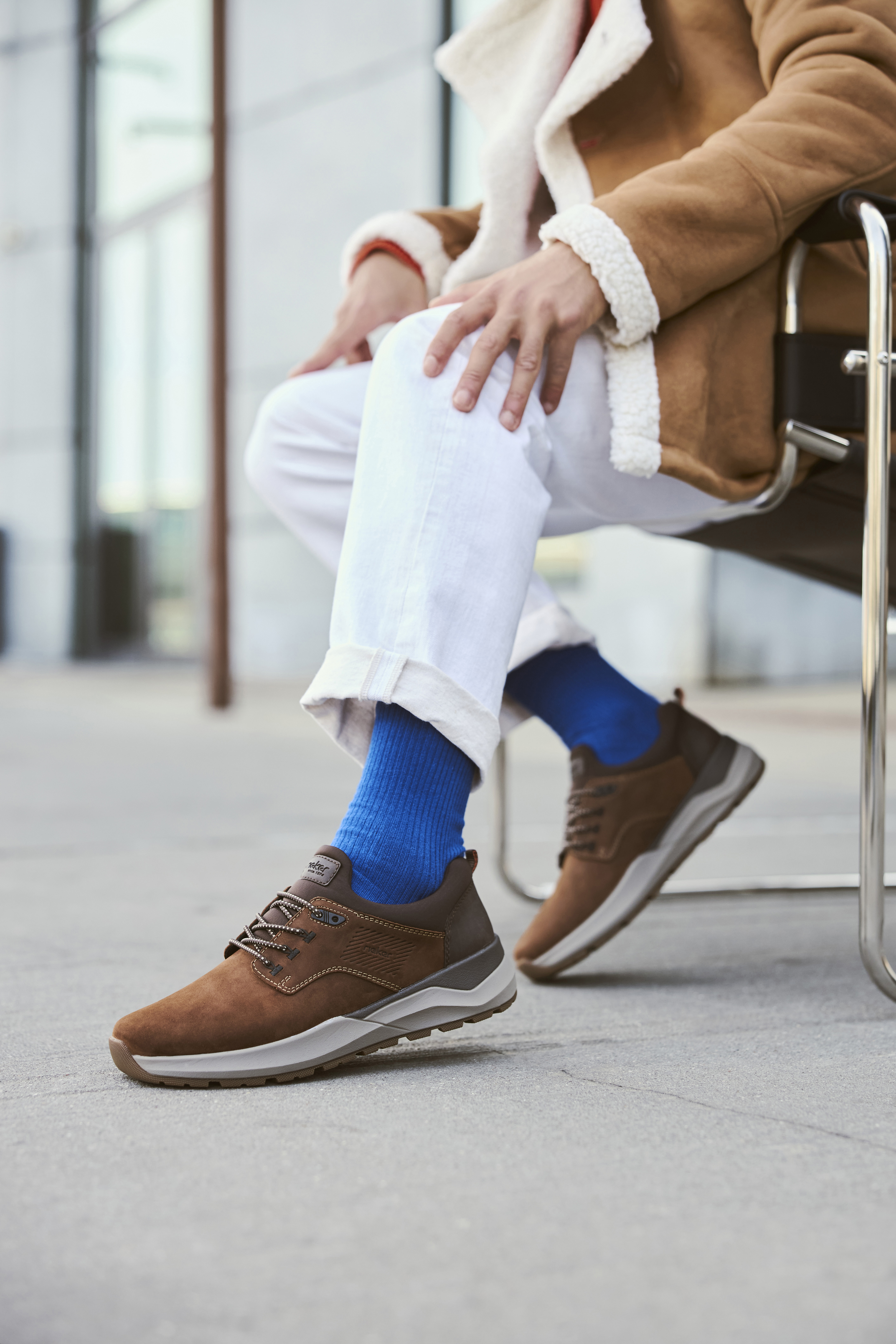 Man draagt bruine Rieker herensneakers met grijze zool en subtiele details, gecombineerd met witte broek en blauwe sokken, voor een casual en comfortabele look.