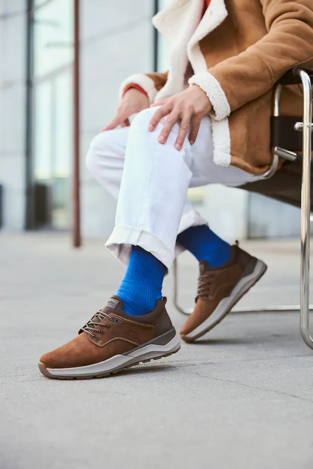 Man draagt bruine Rieker herensneakers met grijze zool en subtiele details, gecombineerd met witte broek en blauwe sokken, voor een casual en comfortabele look.