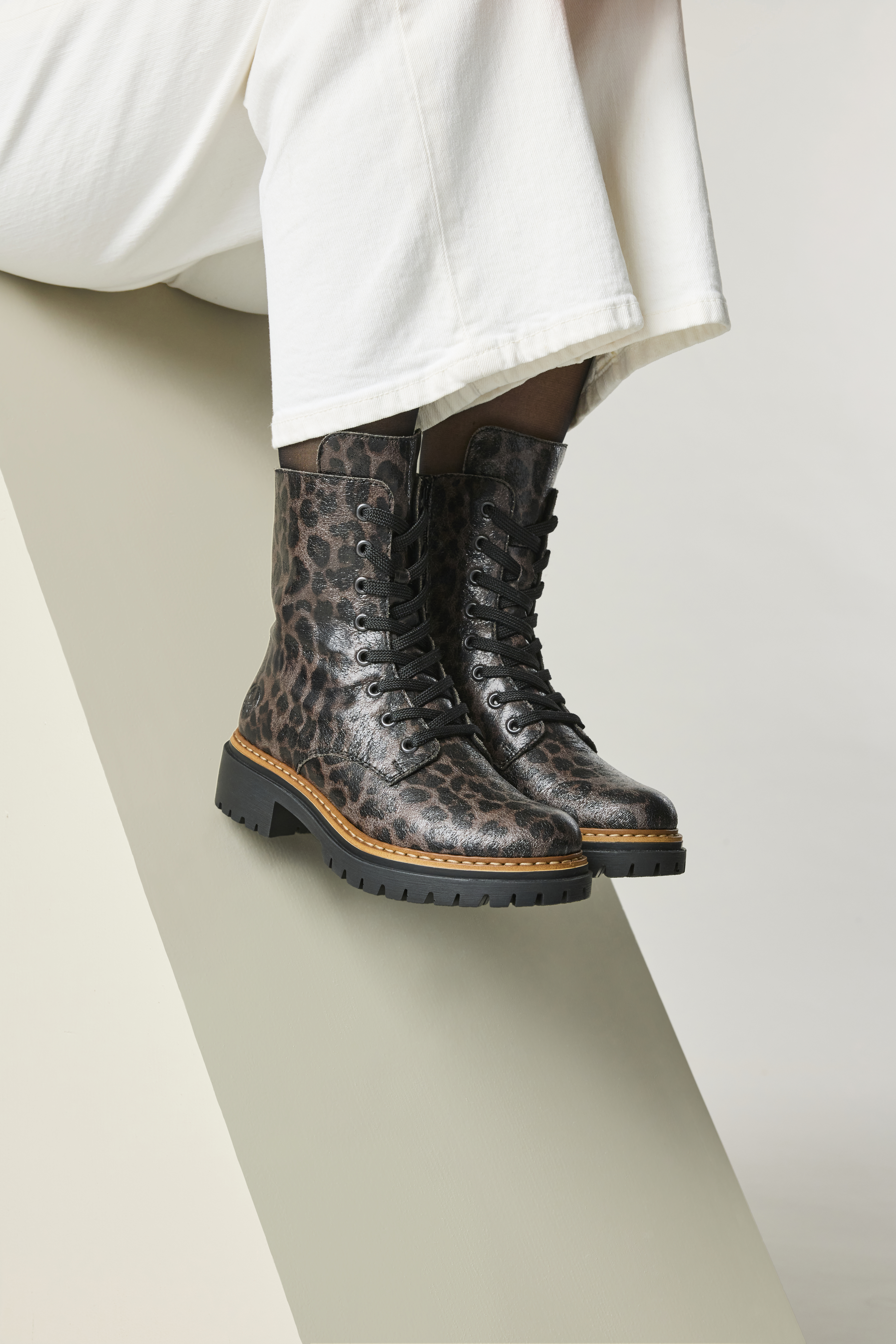 Close-up van Rieker dames veterboots met luipaardprint en zwarte veters, gedragen met witte broek, op een moderne achtergrond.