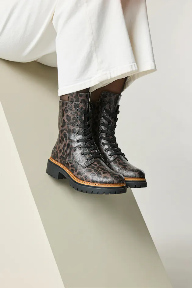 Close-up van Rieker dames veterboots met luipaardprint en zwarte veters, gedragen met witte broek, op een moderne achtergrond.