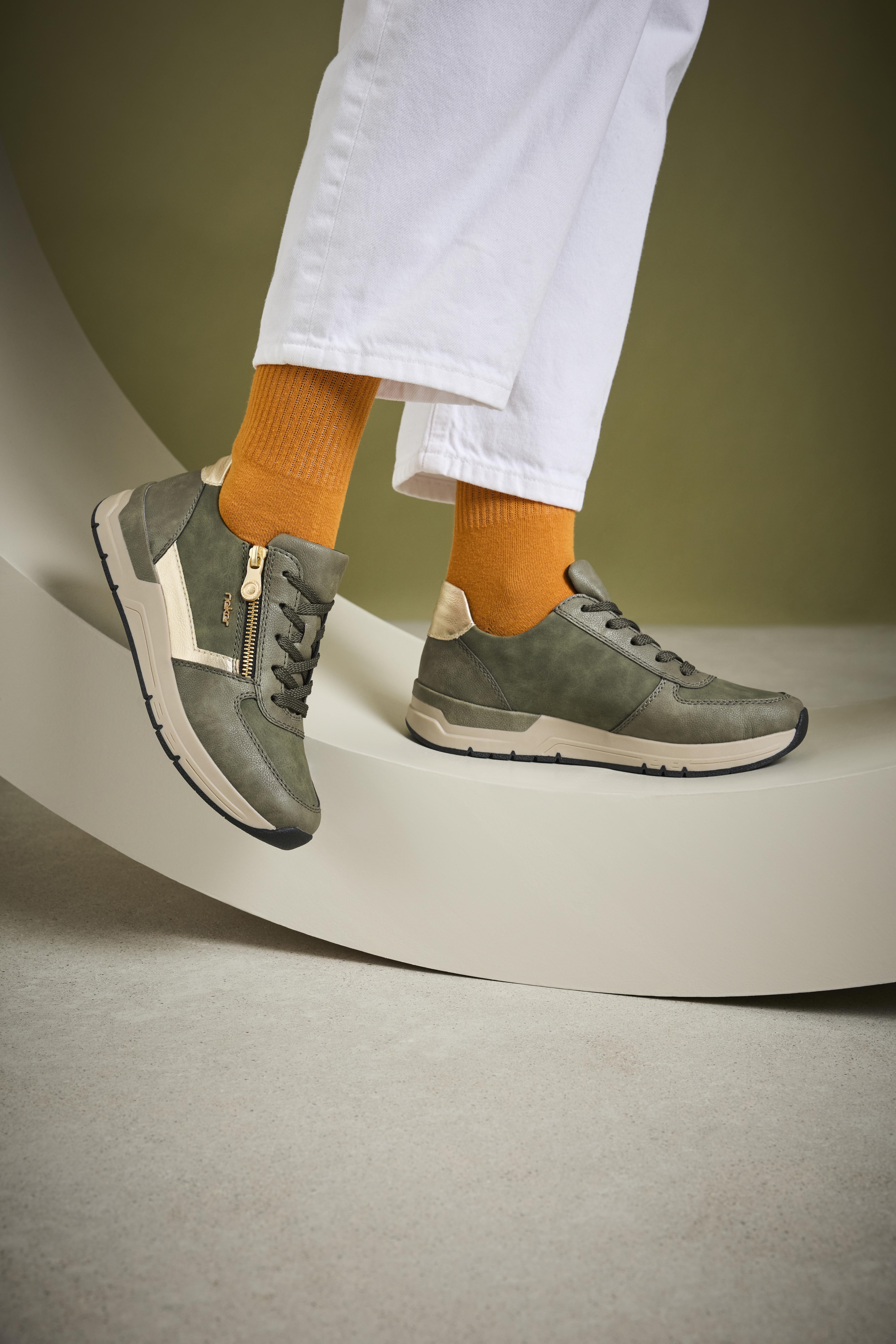 Stijlvolle groene Rieker damessneakers met goudkleurige accenten en rits, gedragen met witte broek en oranje sokken voor een trendy en speelse look.