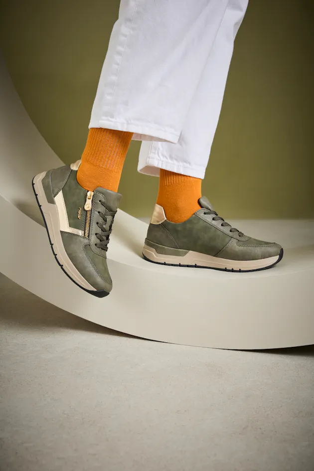 Stijlvolle groene Rieker damessneakers met goudkleurige accenten en rits, gedragen met witte broek en oranje sokken voor een trendy en speelse look.