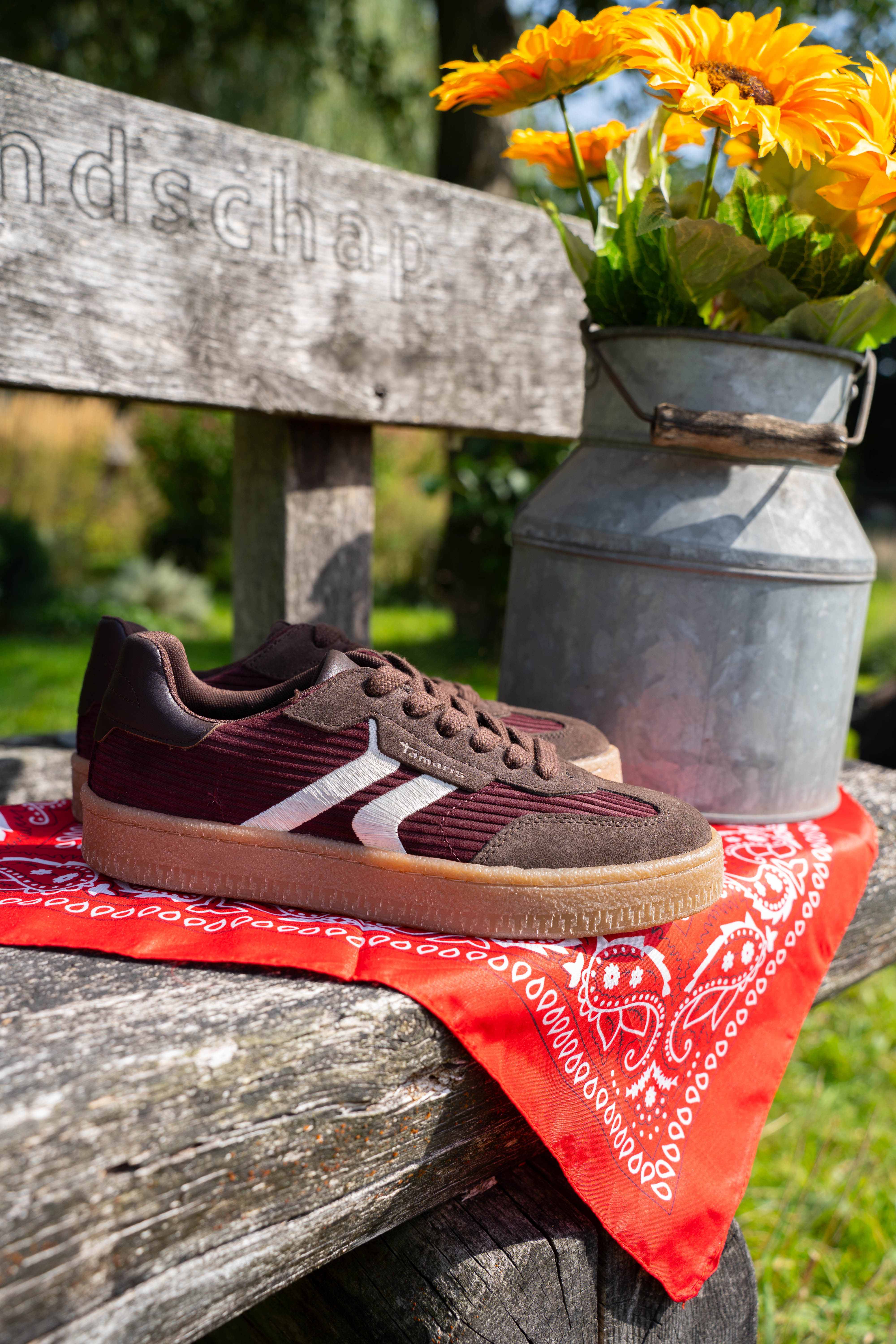 Bruine sneaker met witte details geplaatst op een rood bandana-doek op een houten bankje, naast een melkkan met zonnebloemen.