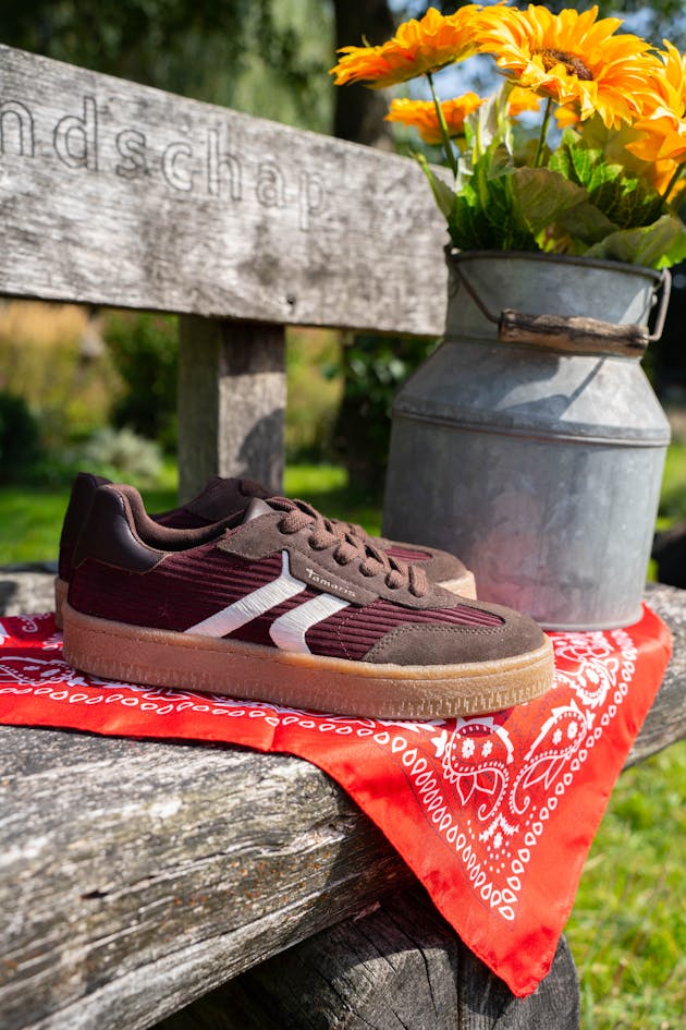 Bruine sneaker met witte details geplaatst op een rood bandana-doek op een houten bankje, naast een melkkan met zonnebloemen.