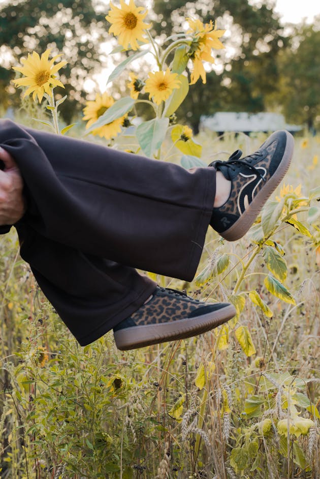 Close-up van damesschoenen in zwart met luipaarddetail, gedragen bij een donkere broek in een veld met zonnebloemen.
