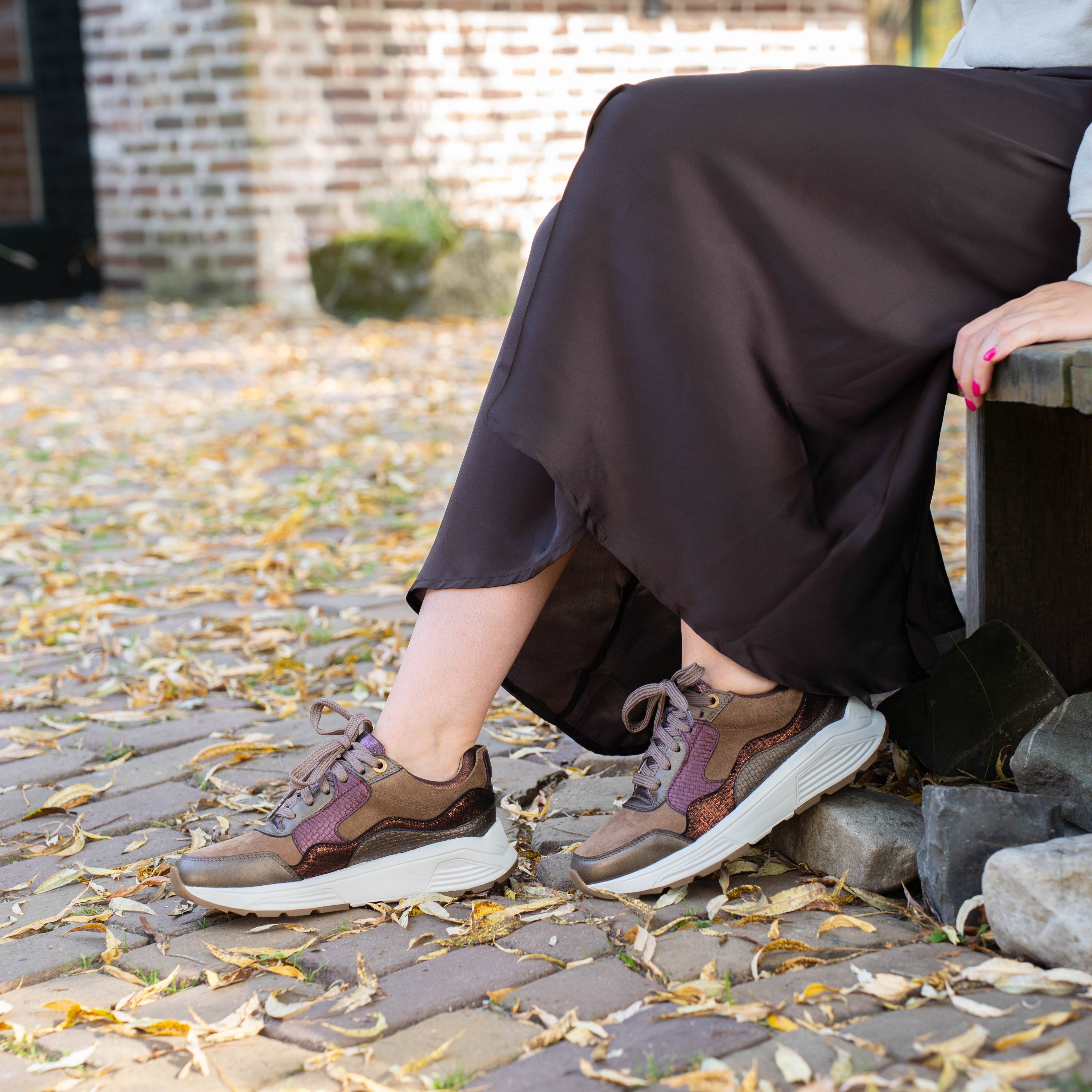 Vrouw met lange rok zittend op een bankje, dragend bruine sneakers met witte zool, omringd door herfstbladeren op straat.