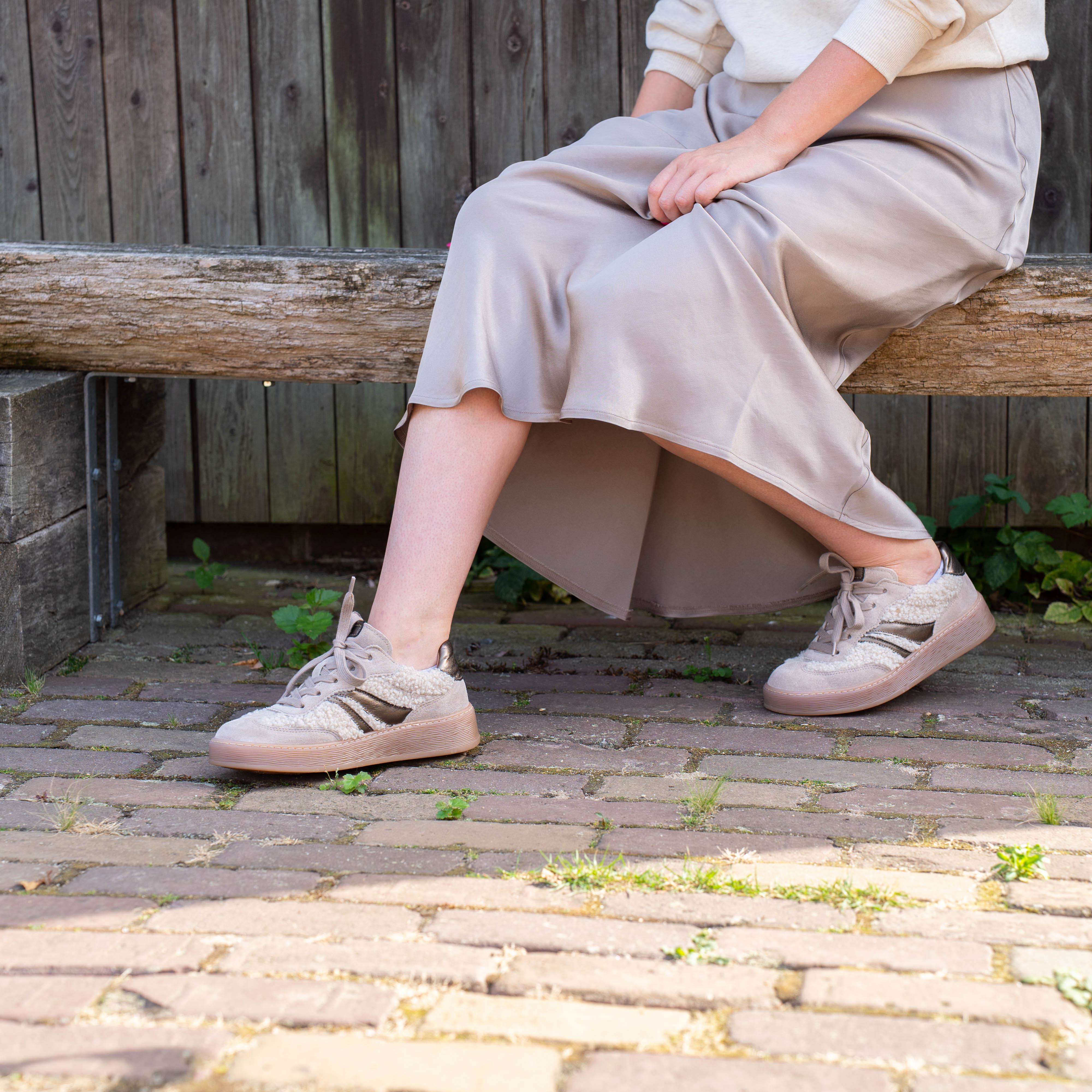 Vrouw met lichte rok zittend op een houten bankje, dragend beige sneakers met witte zool op een zonnige dag.