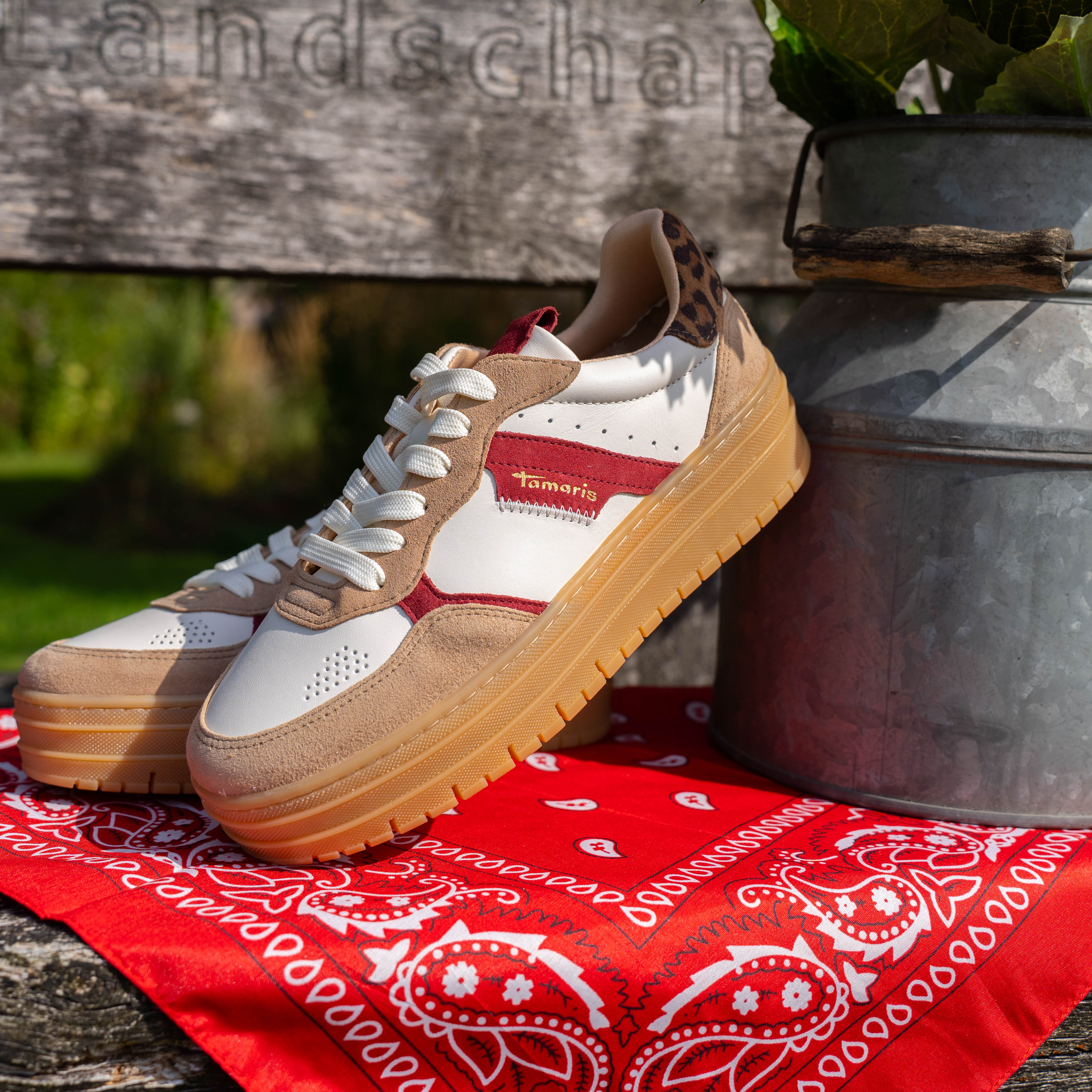 Witte sneakers met rode strepen en dikke houten zool, geplaatst op een rode bandana op een houten bankje naast een melkbus met bloemen.
