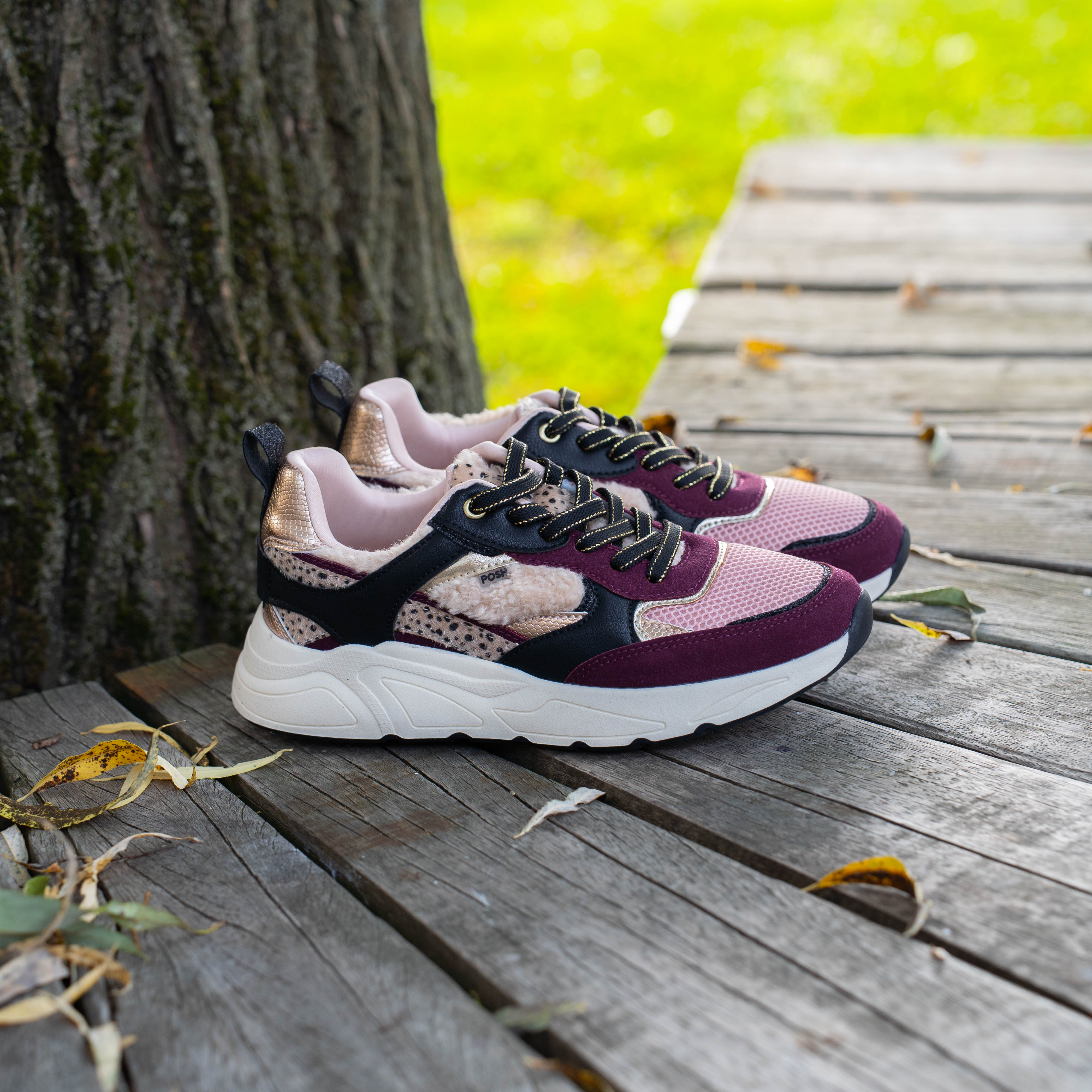 Paars-roze sneakers met witte dikke zool en zwarte details, geplaatst op een houten vlonder naast een boom.