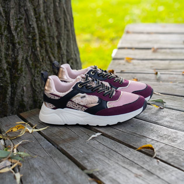 Paars-roze sneakers met witte dikke zool en zwarte details, geplaatst op een houten vlonder naast een boom.