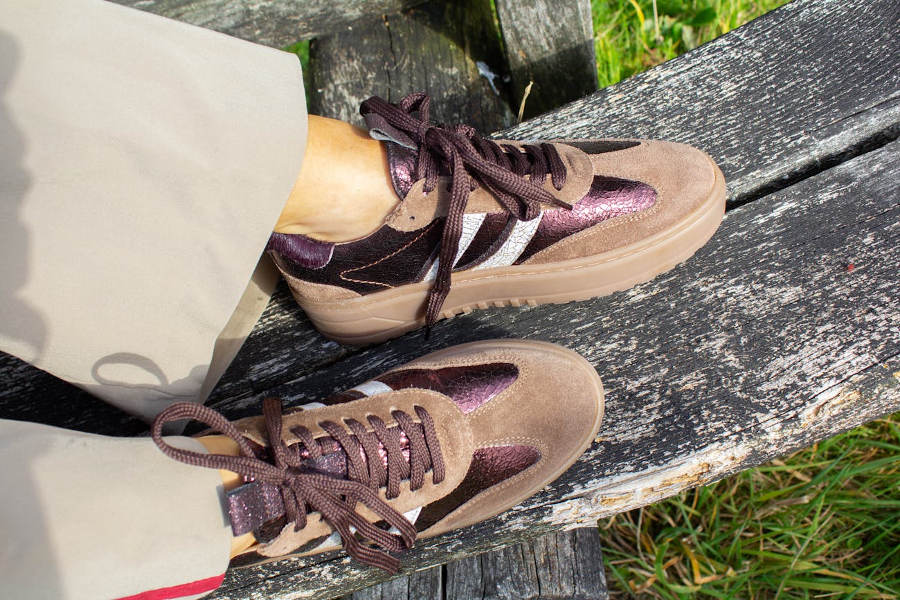 Bruin-paarse sneakers met witte streep en dikke zool, gedragen op een houten bank in het zonlicht.