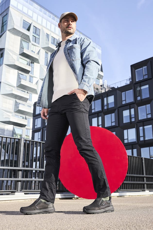 Man draagt zwarte instappers, zwarte broek en een lichte trui met denim jasje, staand voor moderne architectuur en een rode kunstcirkel.