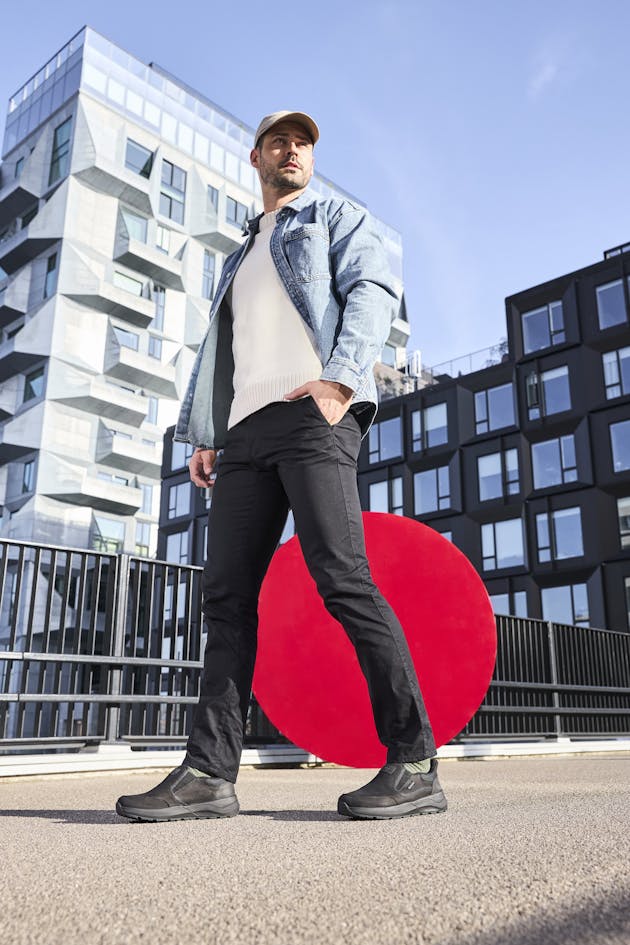 Man draagt zwarte instappers, zwarte broek en een lichte trui met denim jasje, staand voor moderne architectuur en een rode kunstcirkel.