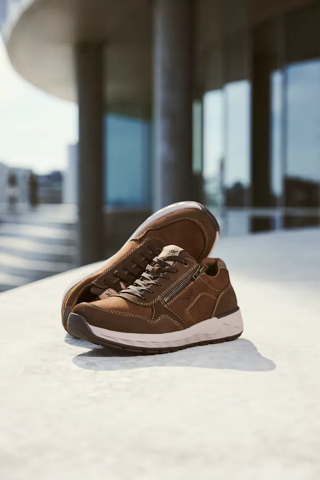 Bruine Rieker heren sneakers van suède met veter en rits, comfortabele zool, gefotografeerd op een moderne achtergrond.