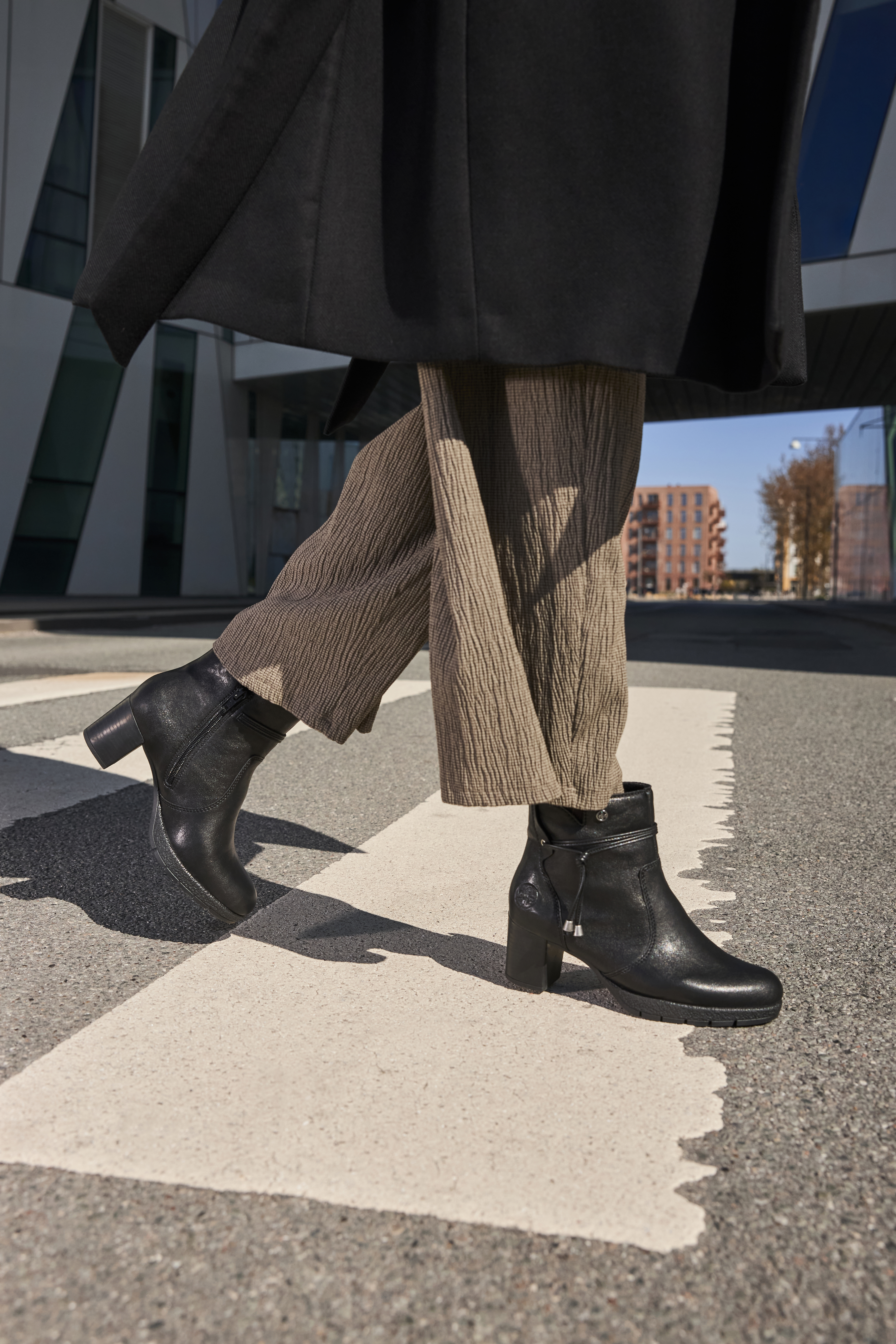 Dames loopt op straat met zwarte enkellaarsjes met hak en rits, gecombineerd met een zwarte jas en wijde broek, voor een stijlvolle en moderne uitstraling.