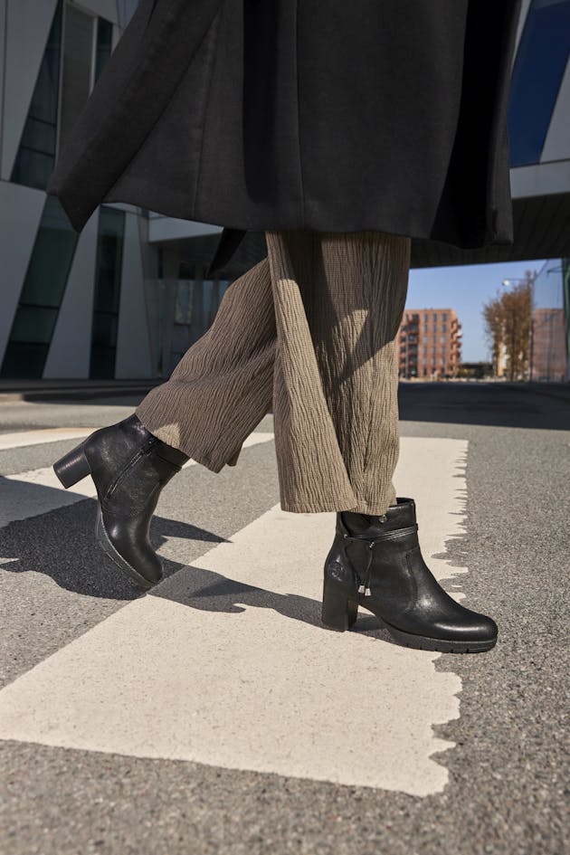 Dames loopt op straat met zwarte enkellaarsjes met hak en rits, gecombineerd met een zwarte jas en wijde broek, voor een stijlvolle en moderne uitstraling.