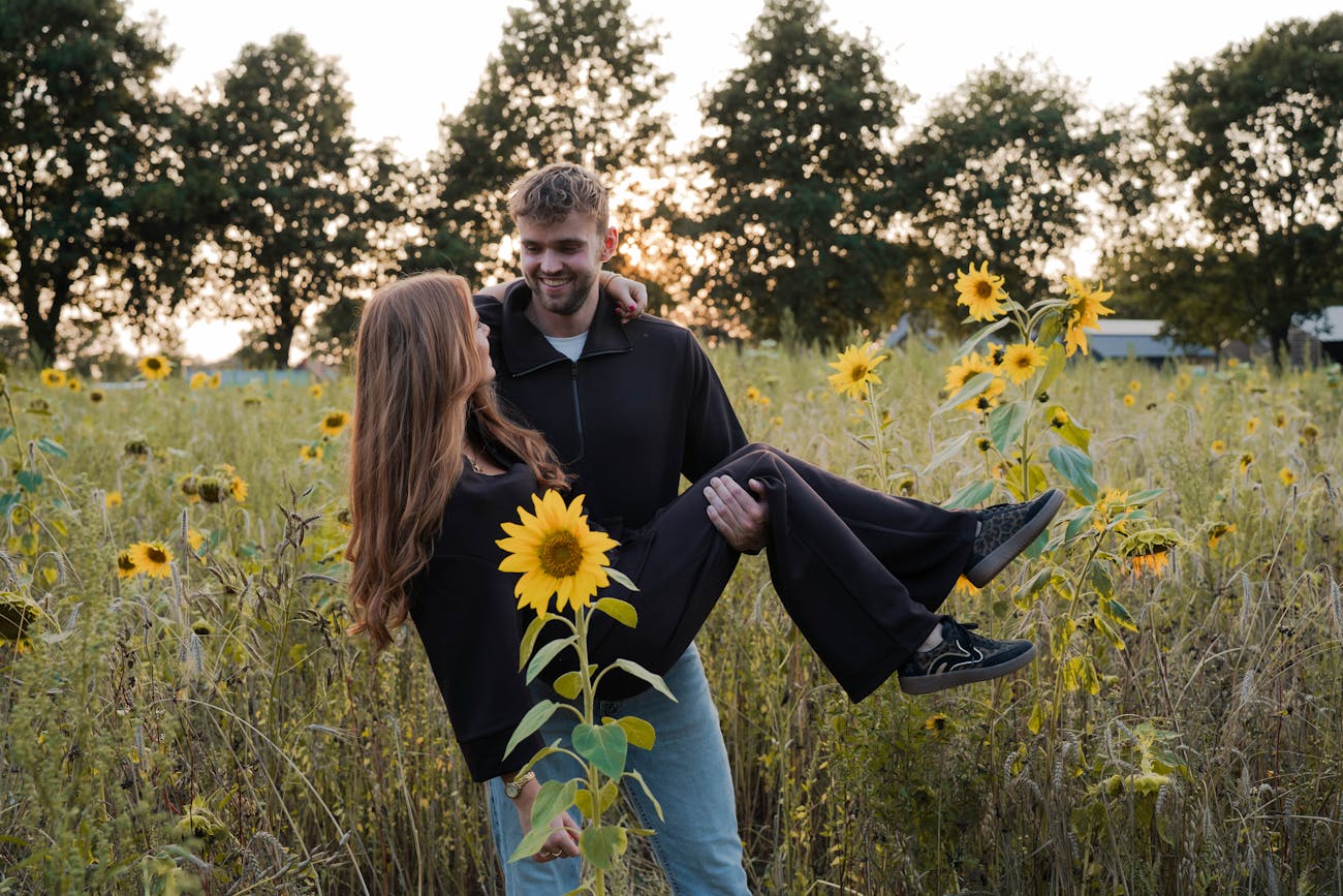 Man tilt vrouw lachend op in een veld vol zonnebloemen tijdens zonsondergang, romantische en warme sfeer.