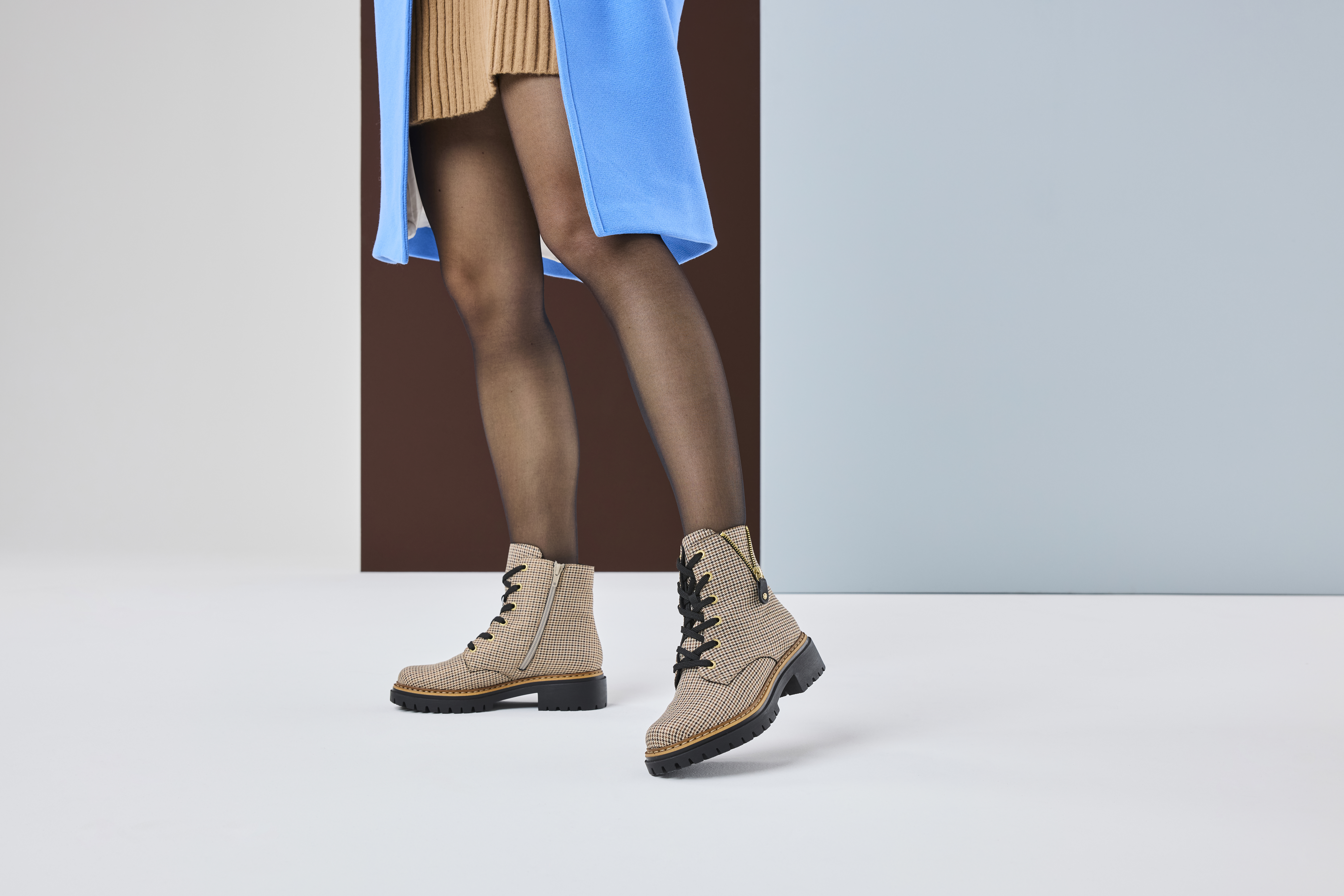 Vrouw draagt beige Rieker veterboots met zwarte veters en stevige zool, gecombineerd met panty en blauwe jas voor een trendy look.