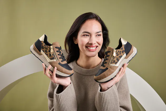 Vrouw houdt een paar Rieker sneakers vast met luipaardprint, beige en zwarte accenten, en lichte zool voor optimaal draagcomfort.