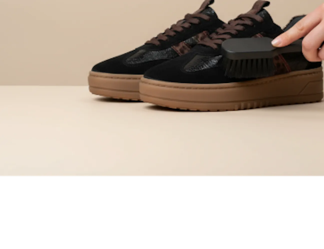 Hand die een suède sneaker met bruine zool voorzichtig schoonborstelt op een beige achtergrond.