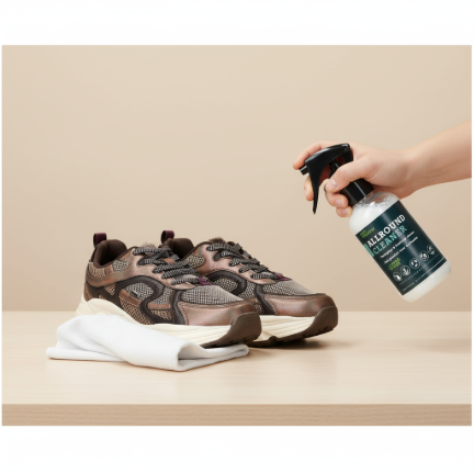 Hand die Allround Cleaner sprayt op een paar bruine sneakers, met een witte doek ernaast op een beige tafel.