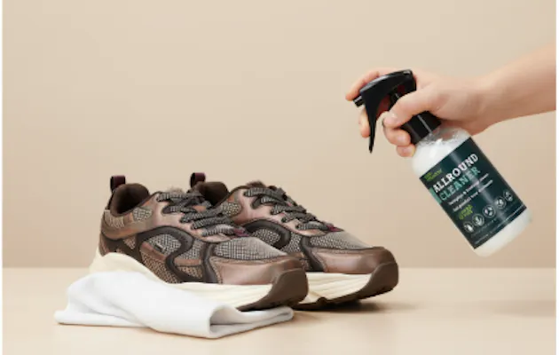 Hand die Allround Cleaner sprayt op een paar bruine sneakers, met een witte doek ernaast op een beige tafel.