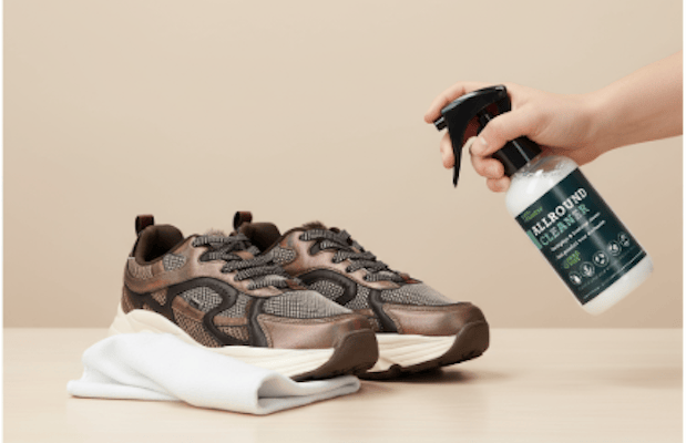 Hand die Allround Cleaner sprayt op een paar bruine sneakers, met een witte doek ernaast op een beige tafel.