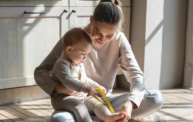 Moeder zit met haar baby op de grond en meet de kleine voet met een geel centimeterlint in een zonnige kamer met warme, natuurlijke tinten.