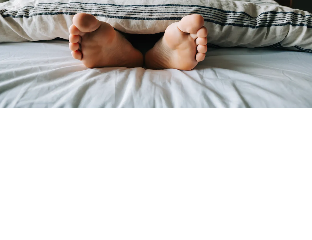 Voeten steken onder het dekbed uit in een warme slaapkamer met twee nachtlampjes, een rustgevende sfeer en zacht ochtendlicht.