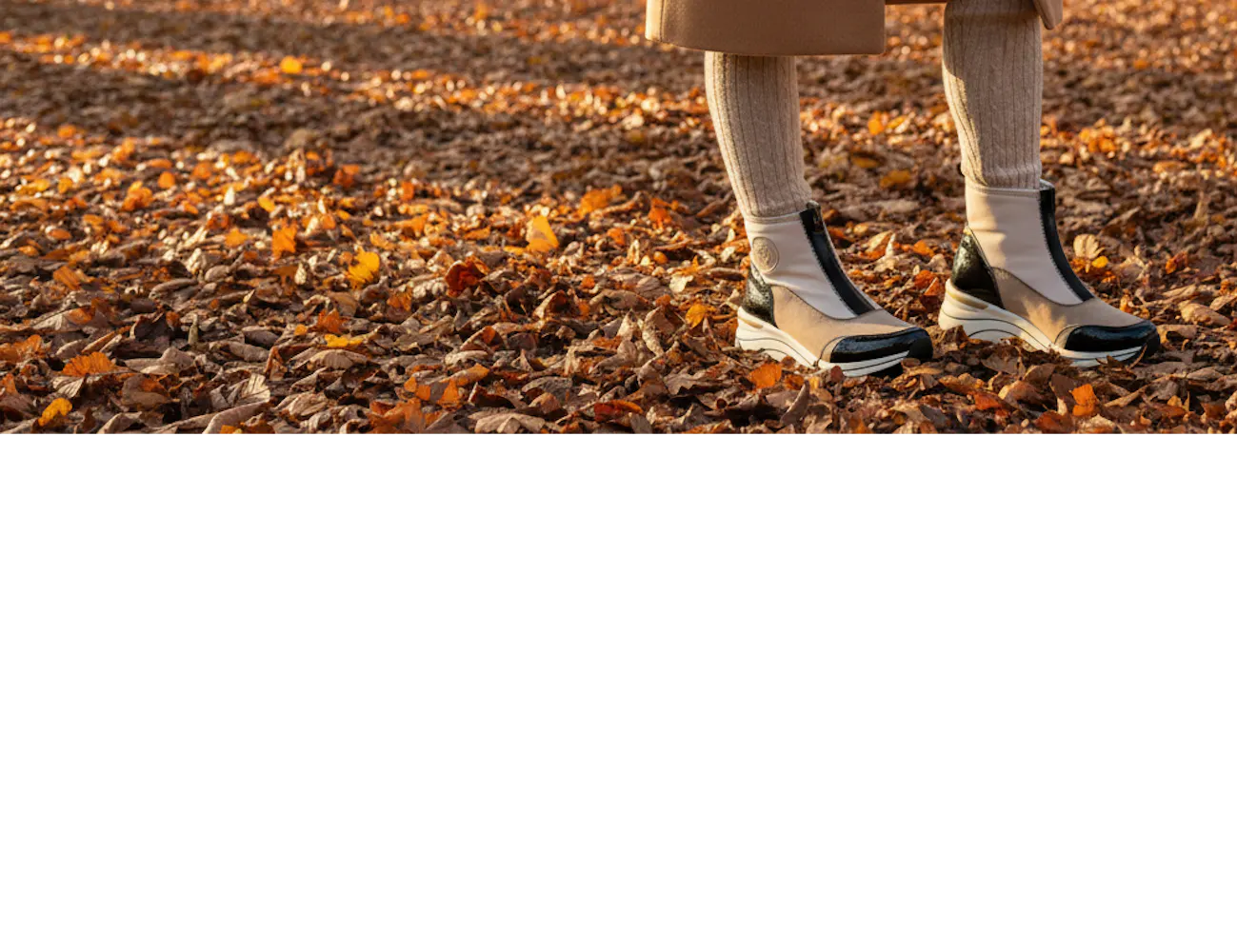 Vrouw die door een park vol herfstbladeren loopt met beige-zwarte chelseaboots, gecombineerd met een lange camel jas en warme knitwear.