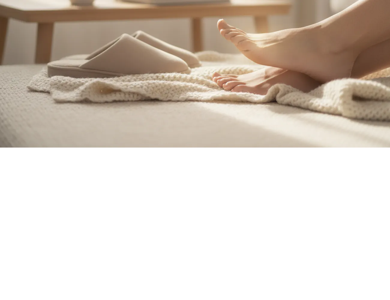 Blote voeten rustend op een plaid naast beige pantoffels, met een kop dampende koffie op een houten tafeltje in een zachte, lichte woonkamer.