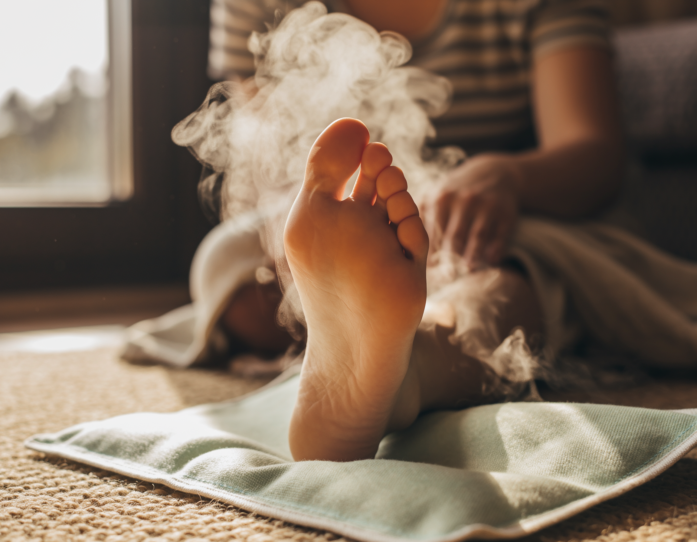 Close-up van een voet die rust op een warmtekussen, met opstijgende stoom en warm zonlicht voor een ontspannen, huiselijke sfeer.