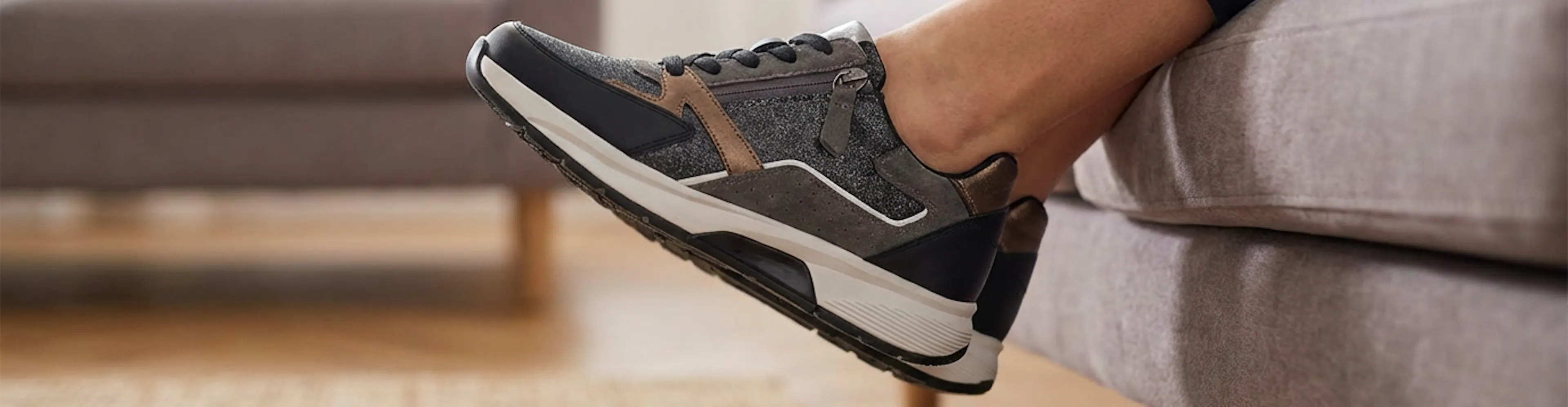 Vrouwelijk been met grijs-bruine sneaker met rits, ontspannen omhoog gehouden terwijl ze op een bank zit in een warme, huiselijke woonkamer.