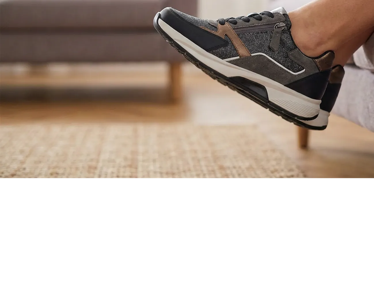 Vrouwelijk been met grijs-bruine sneaker met rits, ontspannen omhoog gehouden terwijl ze op een bank zit in een warme, huiselijke woonkamer.