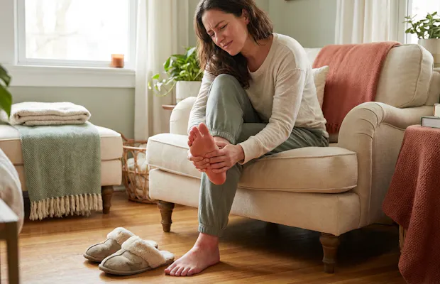Vrouw die haar voet vasthoudt vanwege pijn terwijl ze op een beige fauteuil zit in een warme, huiselijke woonkamer met sloffen op de vloer.