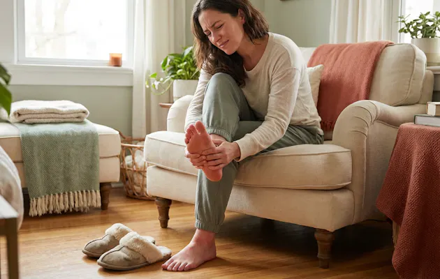 Vrouw die haar voet vasthoudt vanwege pijn terwijl ze op een beige fauteuil zit in een warme, huiselijke woonkamer met sloffen op de vloer.